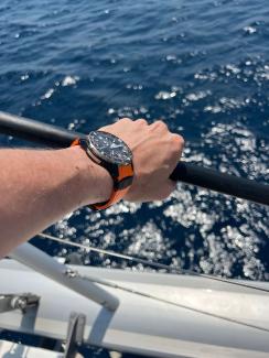 Unterarm mit orange-schwarzer Uhr, die Hand hält sich an der Reling eines Schiffes auf Wasser fest